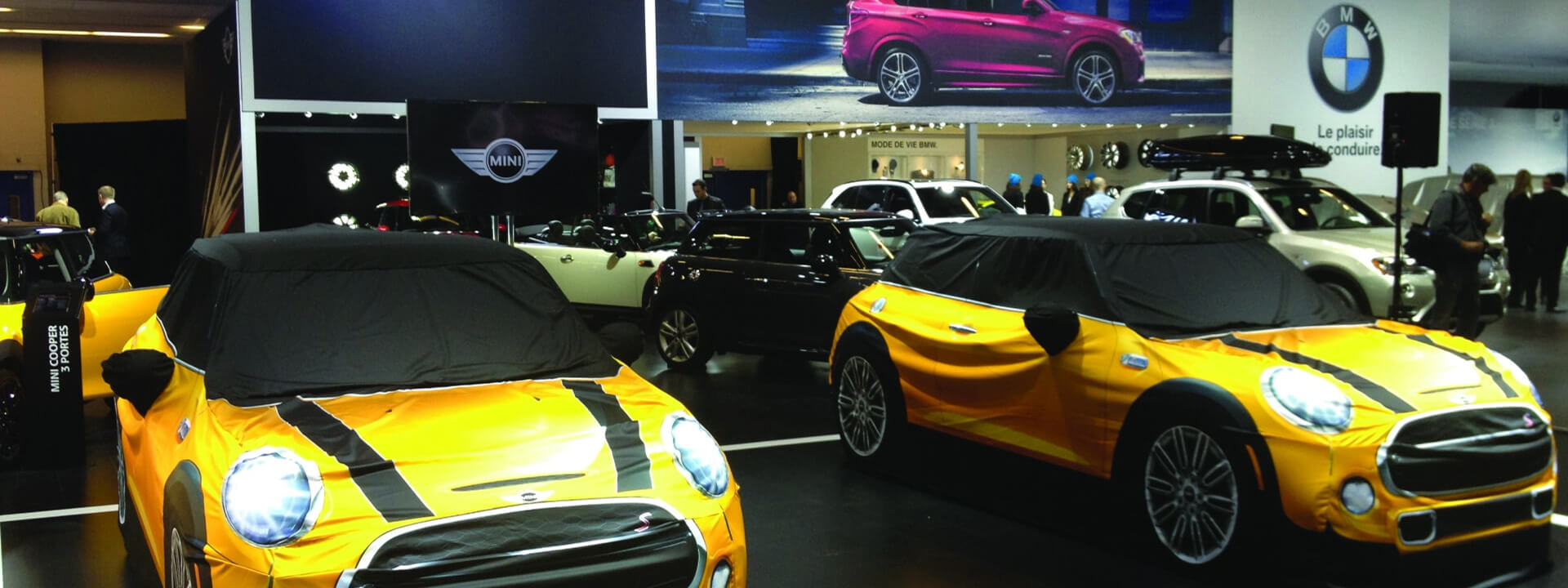 "Two yellow Mini Coopers, covered in graphic fabrics depicting their features and racing stripes, are displayed at a car show with other BMW and Mini vehicles in the background."