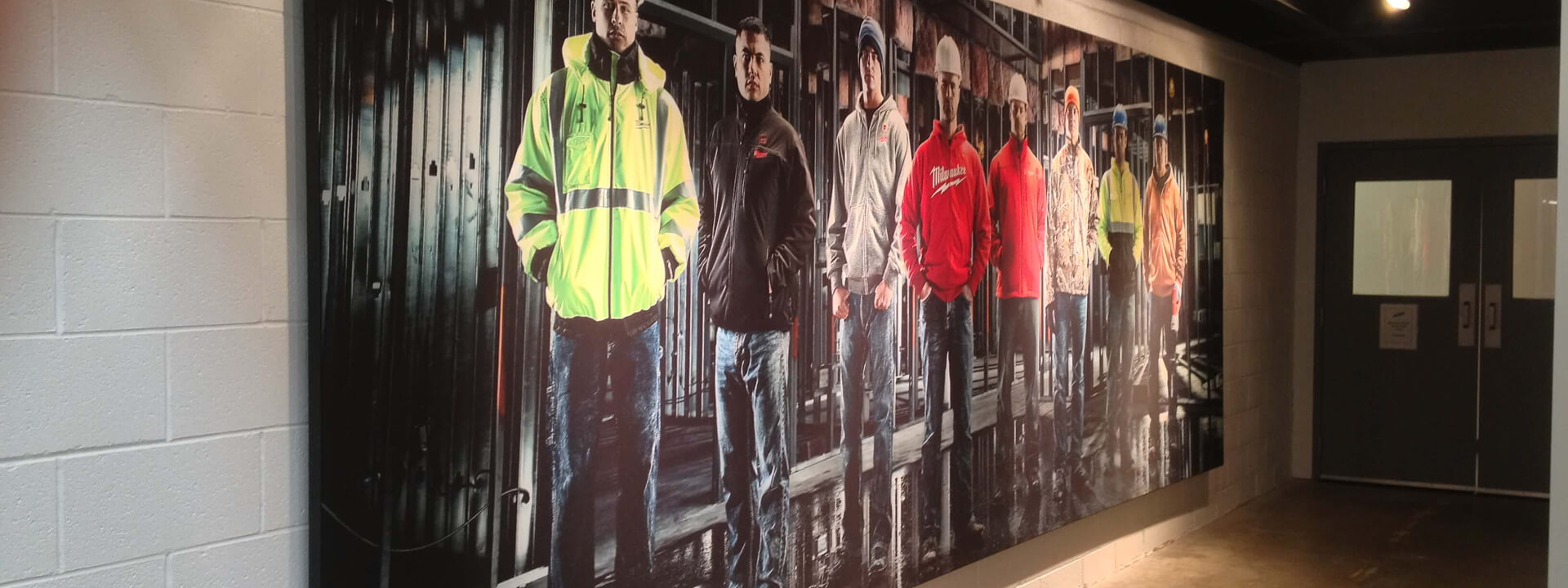 A large wall mural in a hallway shows a lineup of men wearing construction workwear, some with Milwaukee Tool branding.