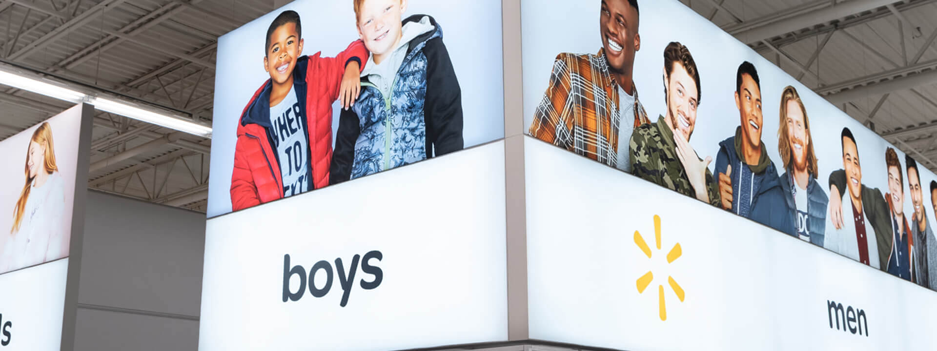Overhead illuminated signs in a Walmart store display smiling models for 'boys' and 'men' clothing sections, featuring the spark logo.