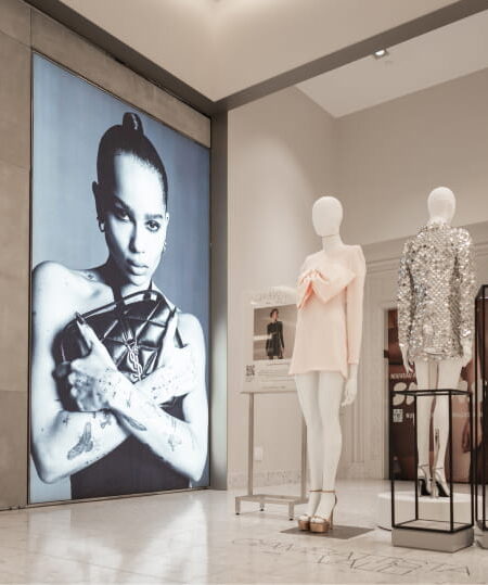 "A YSL fashion display features a large black and white photo of Zoë Kravitz holding a YSL bag, a glowing YSL logo, and two mannequins in a pink bow dress and a silver sequin outfit."