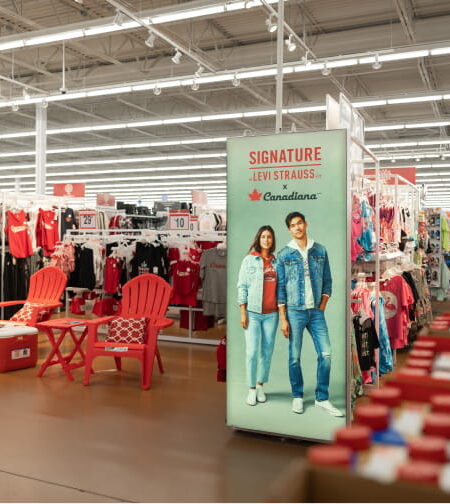 A department store aisle with clothing racks, a large ""Signature Levi Strauss x Canadiana"" ad banner, and red patio chairs on display.