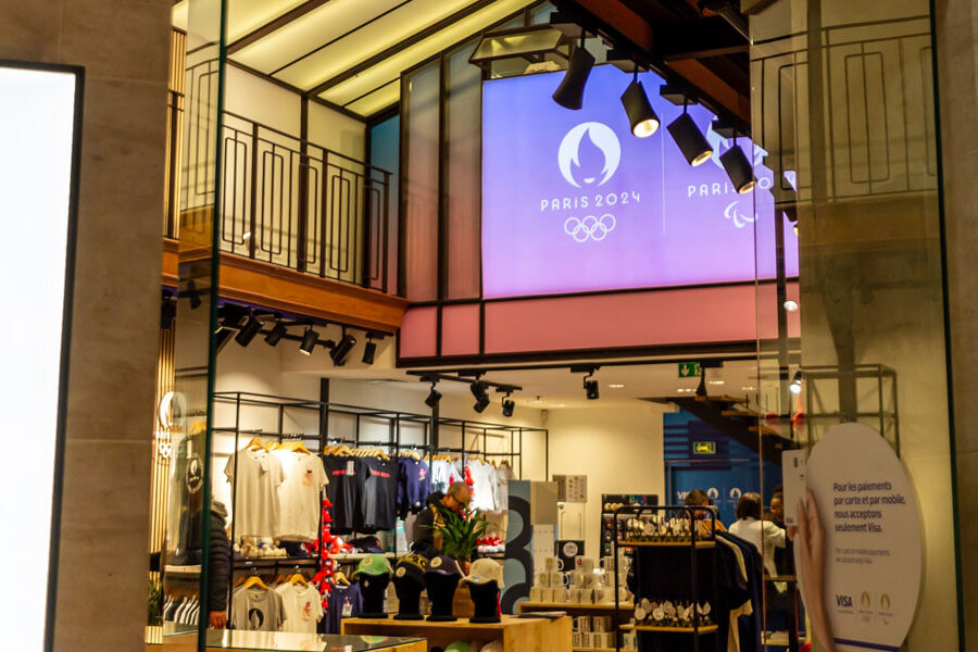 A Paris 2024 merchandise store featuring branded apparel and a large screen displaying the event logo and Olympic rings.