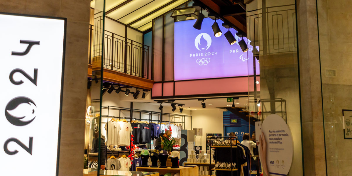 A Paris 2024 merchandise store featuring branded apparel and a large screen displaying the event logo and Olympic rings.