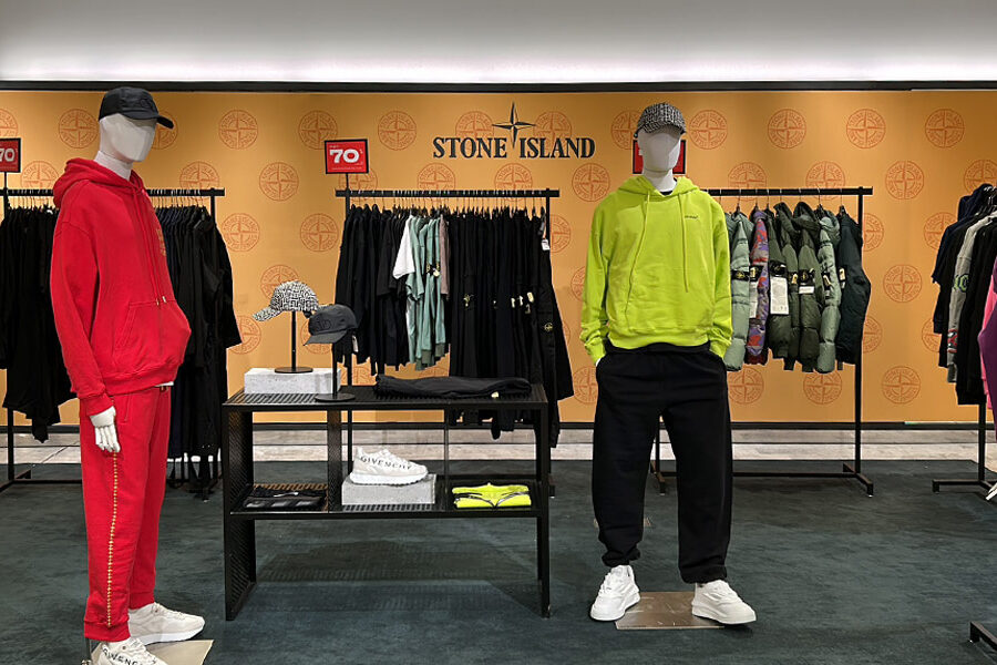 Stone Island retail display featuring two mannequins in a red tracksuit and a neon yellow hoodie, against a branded wall with racks of clothing and accessories.