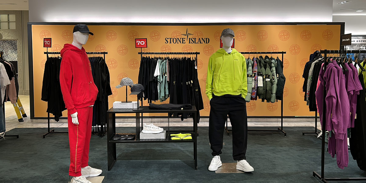 Stone Island retail display featuring two mannequins in a red tracksuit and a neon yellow hoodie, against a branded wall with racks of clothing and accessories.
