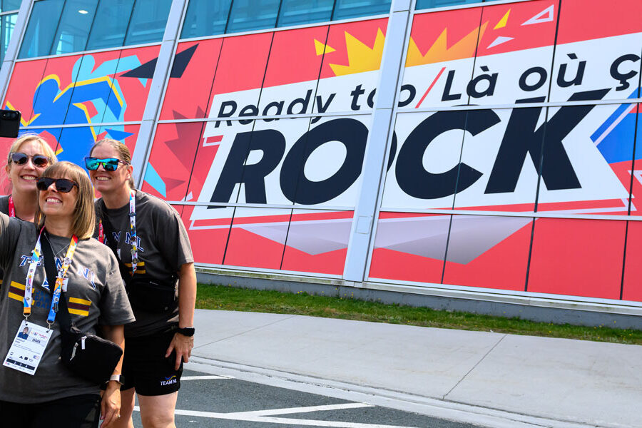 Three smiling women take a selfie in front of a building displaying a large red banner that reads ""Ready to / Là où ça ROCK.""