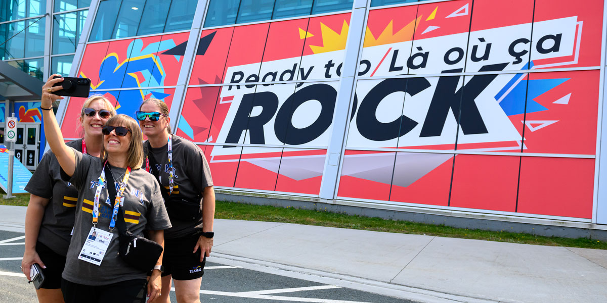 Three smiling women take a selfie in front of a building displaying a large red banner that reads ""Ready to / Là où ça ROCK.""