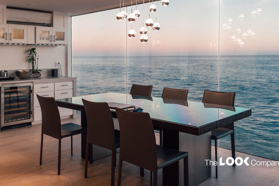 A modern dining room with a glass table, brown chairs, wet bar, and a floor-to-ceiling window overlooking the ocean.