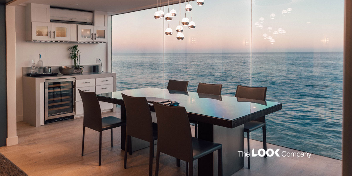 A modern dining room with a glass table, brown chairs, wet bar, and a floor-to-ceiling window overlooking the ocean.