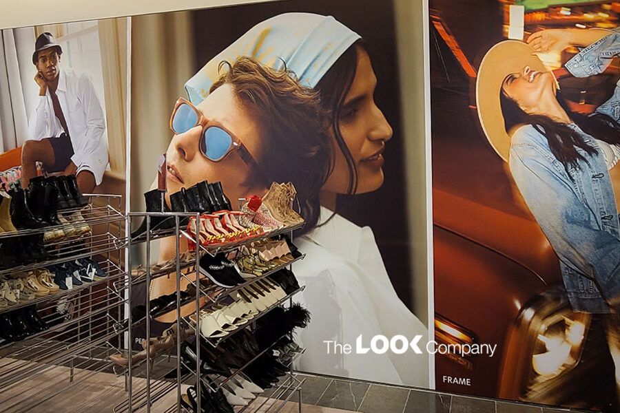 Metal shoe racks displaying various footwear are positioned in front of large wall advertisements featuring fashion models.