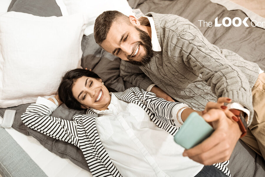 A happy couple smiles while taking a selfie together on a bed.