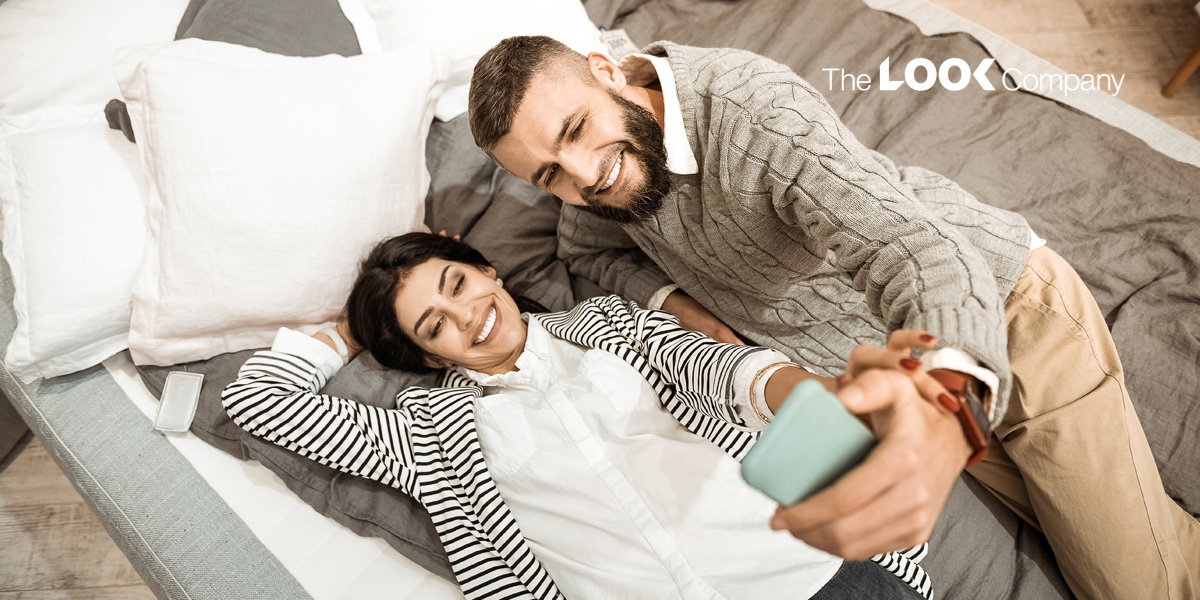A happy couple smiles while taking a selfie together on a bed.
