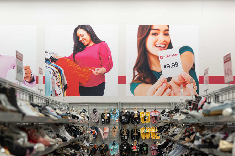 "Shelves full of shoes and sandals line the floor of a Burlington store, below wall advertisements showing happy shoppers and a prominent $9.99 price tag."