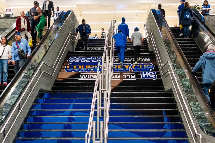 "People ascend and descend escalators flanking a central staircase decorated with ""Coupe Grey Cup Winnipeg 112 HQ"" branding."