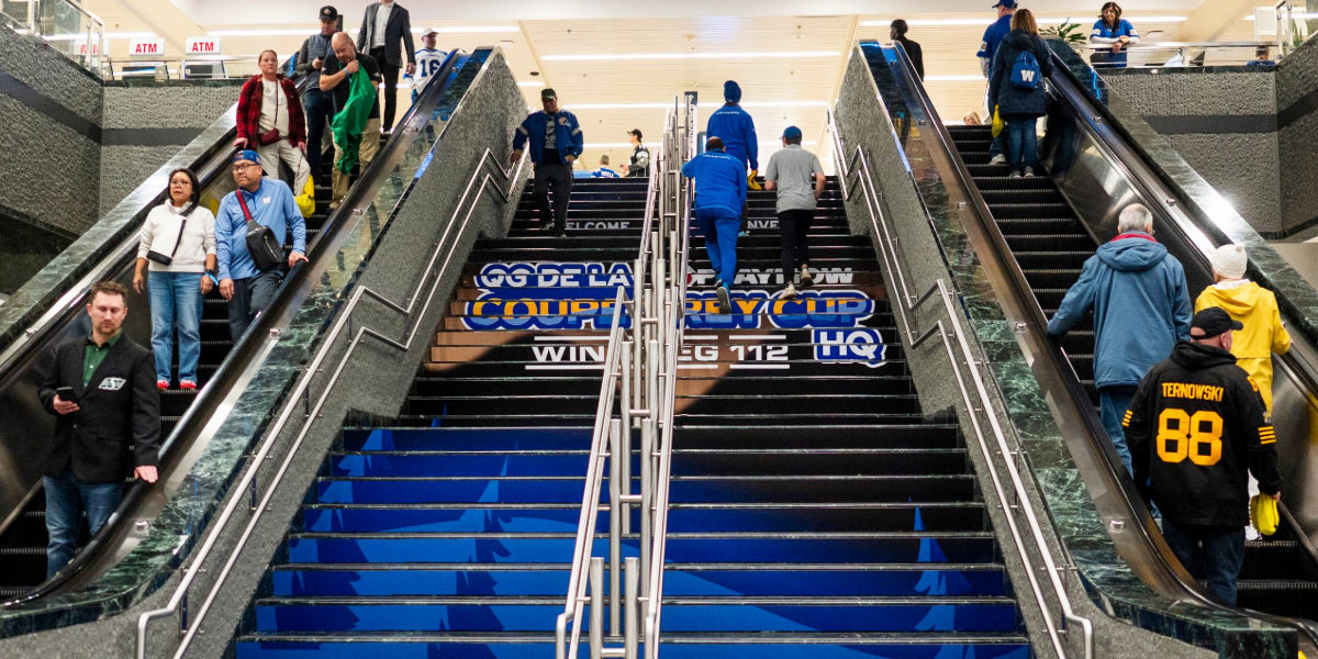 "People ascend and descend escalators flanking a central staircase decorated with ""Coupe Grey Cup Winnipeg 112 HQ"" branding."