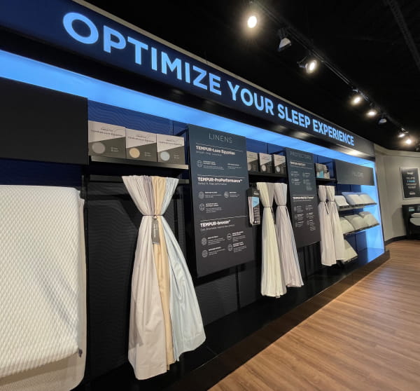 A retail store display features various bedding linens and protectors beneath a sign that reads ""OPTIMIZE YOUR SLEEP EXPERIENCE.""