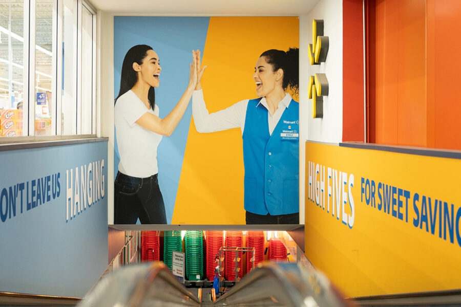 An escalator descends into a Walmart store past a large mural of two women high-fiving, with promotional text ""Don't Leave Us Hanging"" and ""High Fives for Sweet Savings"" on the surrounding walls.