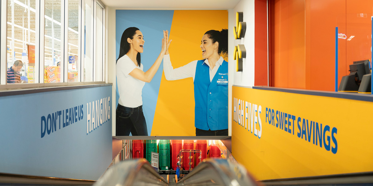 An escalator descends into a Walmart store past a large mural of two women high-fiving, with promotional text ""Don't Leave Us Hanging"" and ""High Fives for Sweet Savings"" on the surrounding walls.
