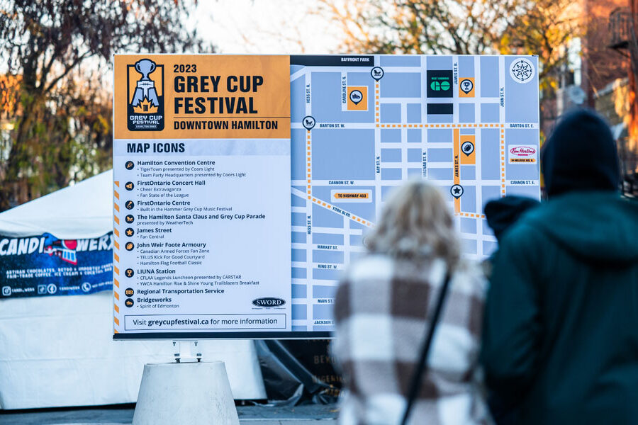 A large outdoor sign displays the map and event information for the 2023 Grey Cup Festival in Downtown Hamilton, with several people nearby on a city street.