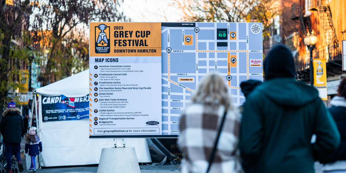 A large outdoor sign displays the map and event information for the 2023 Grey Cup Festival in Downtown Hamilton, with several people nearby on a city street.