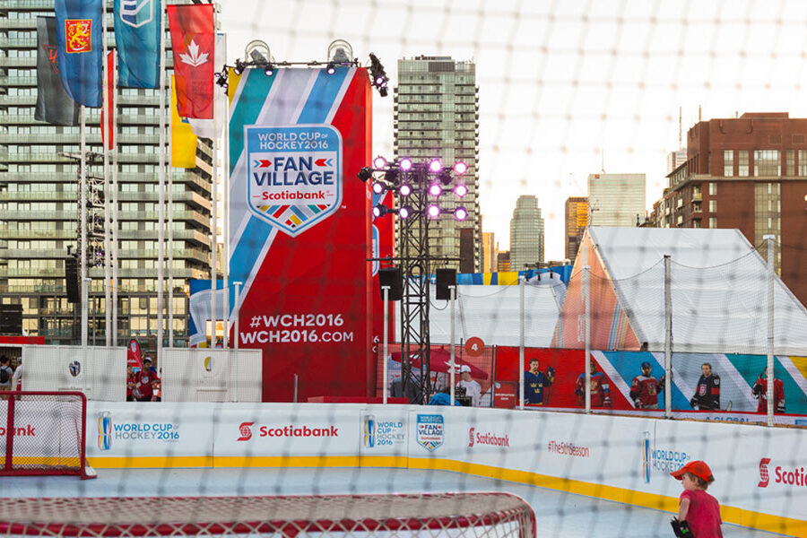 An outdoor hockey rink set up as part of the World Cup of Hockey 2016 Fan Village, featuring large event banners, sponsor branding, and city buildings in the background.