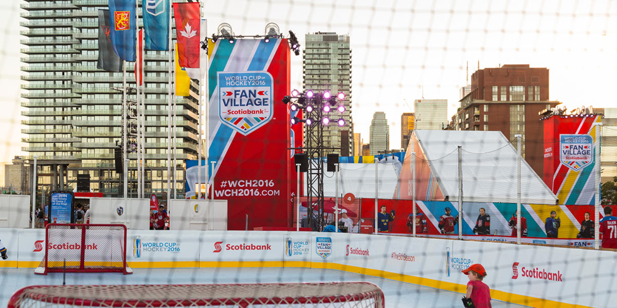 An outdoor hockey rink set up as part of the World Cup of Hockey 2016 Fan Village, featuring large event banners, sponsor branding, and city buildings in the background.