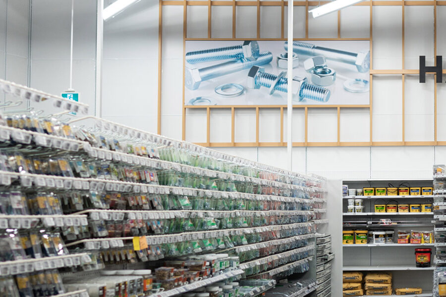 A hardware store aisle displays shelves of packaged fasteners with a large wall graphic of bolts and nuts in the background.