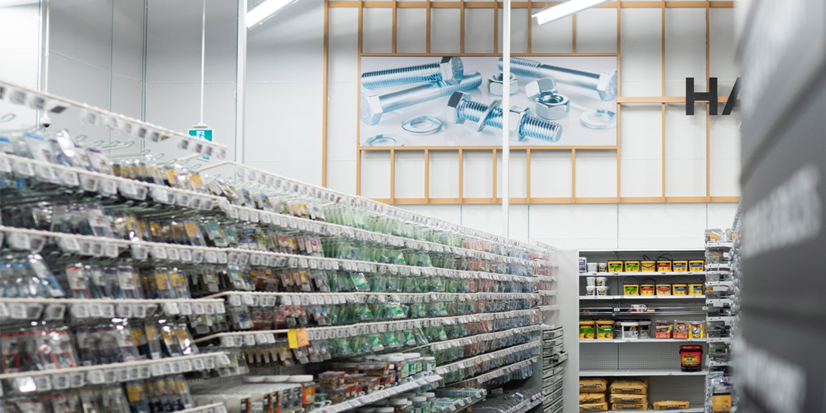 A hardware store aisle displays shelves of packaged fasteners with a large wall graphic of bolts and nuts in the background.