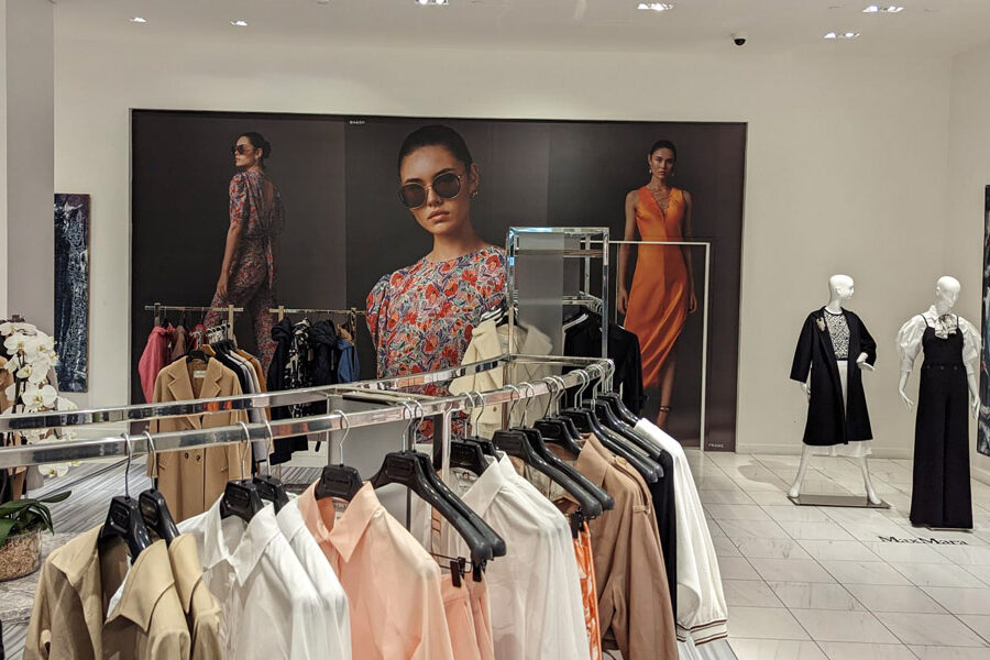 "A brightly lit fashion store interior features racks of clothing, mannequins displaying outfits, and large wall graphics with models and an abstract painting."