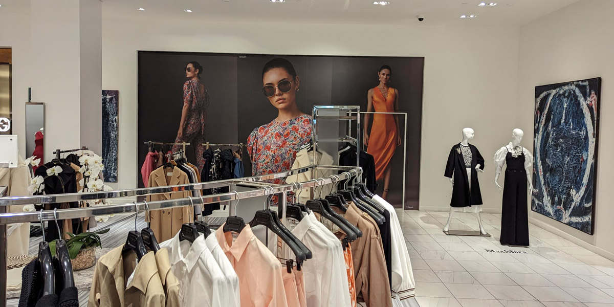 "A brightly lit fashion store interior features racks of clothing, mannequins displaying outfits, and large wall graphics with models and an abstract painting."