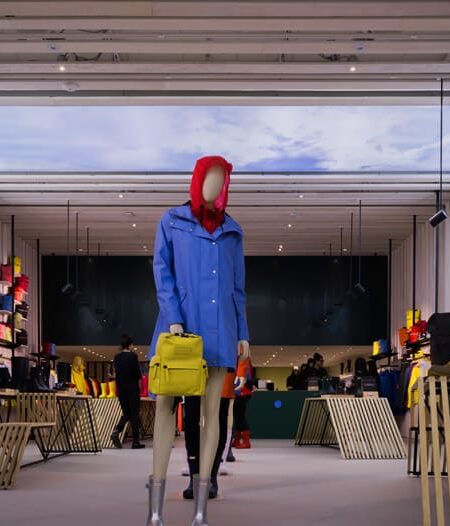 A bright retail store interior features a mannequin in a blue raincoat and yellow backpack, surrounded by clothing displays and wooden tables, with a large sky scene on the back wall.