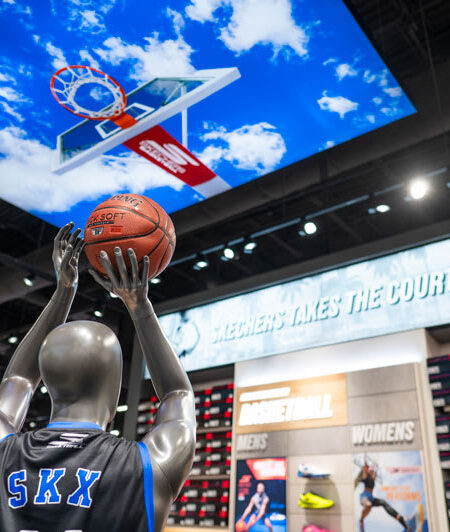 A mannequin in a Skechers basketball jersey aims a ball at a digital ceiling display showing a hoop, with a ""Skechers Takes The Court"" sign in the background.