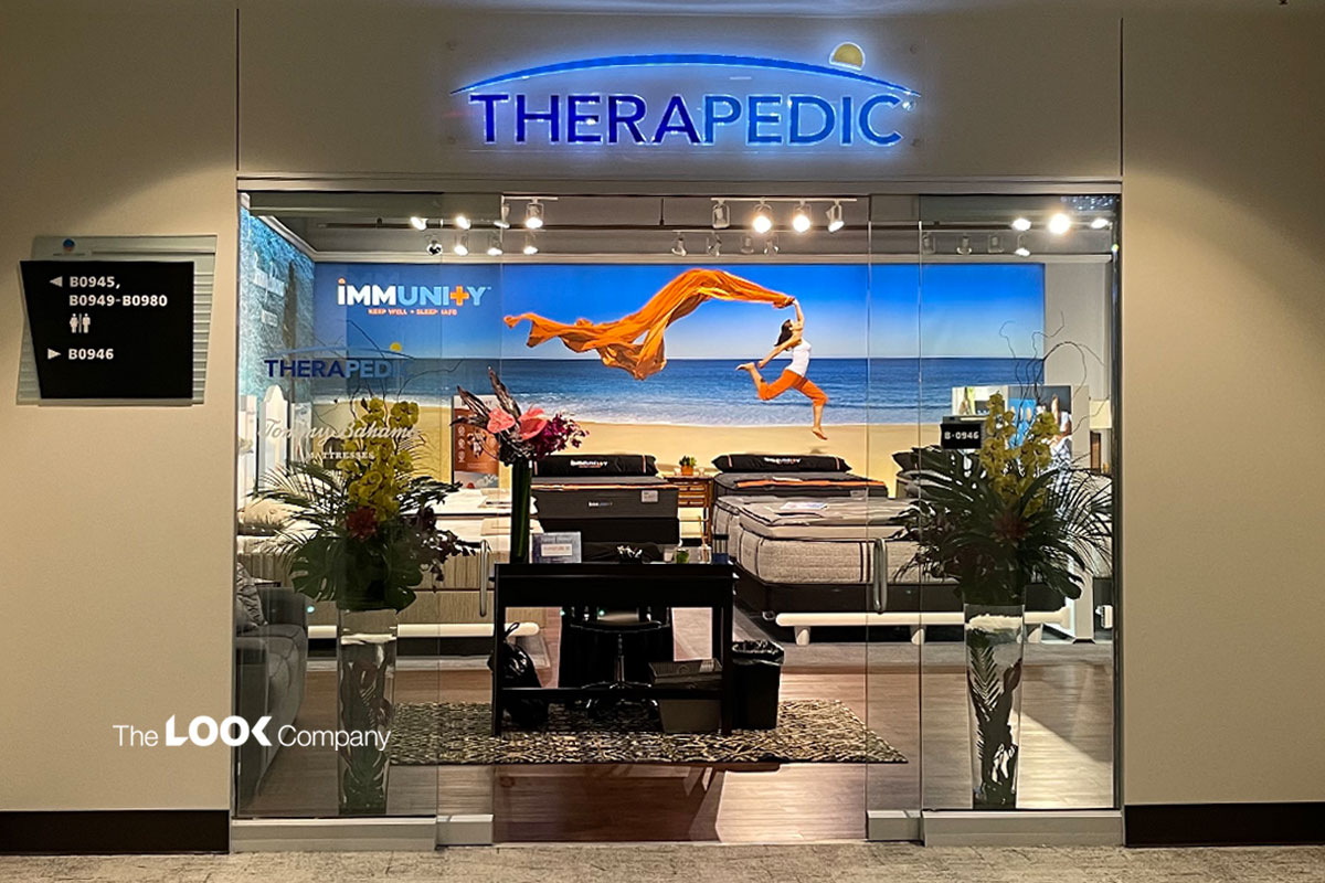 A Therapedic mattress store entrance features a vibrant beach-themed display with a person leaping, and mattresses inside.