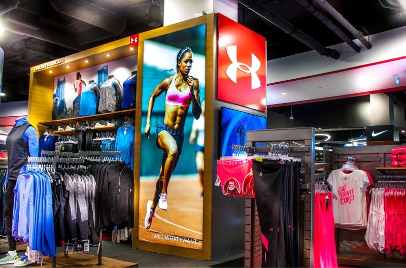 Interior of an Under Armour athletic wear store featuring a large illuminated display of a female runner, branding, and clothing racks.
