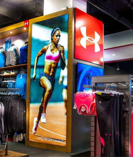 Interior of an Under Armour sports apparel store, featuring a large lit display of a female runner, racks of athletic clothing, and mannequins, with a Nike logo visible in the background.