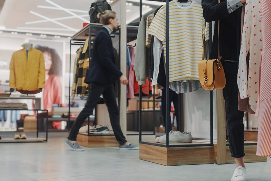 The interior of a contemporary fashion boutique. In the foreground, a person in white sneakers and a tan crossbody bag stands near a rack of striped and patterned dresses. In the blurred background, another shopper walks past minimalist wooden and metal clothing displays under bright, geometric ceiling lights.