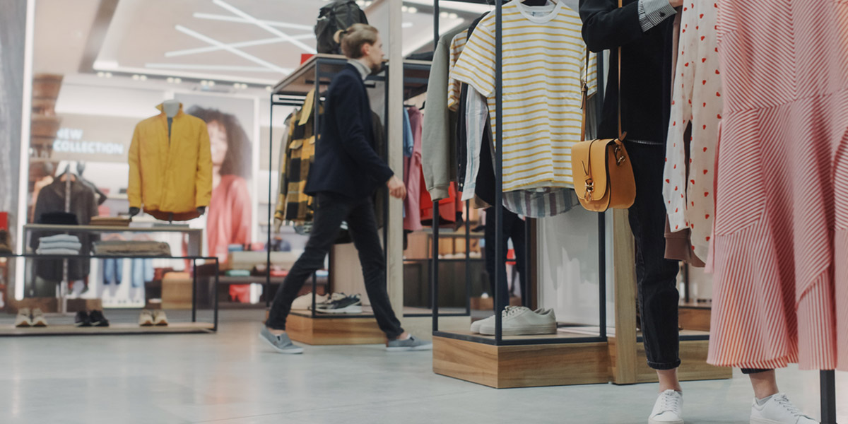 The interior of a contemporary fashion boutique. In the foreground, a person in white sneakers and a tan crossbody bag stands near a rack of striped and patterned dresses. In the blurred background, another shopper walks past minimalist wooden and metal clothing displays under bright, geometric ceiling lights.