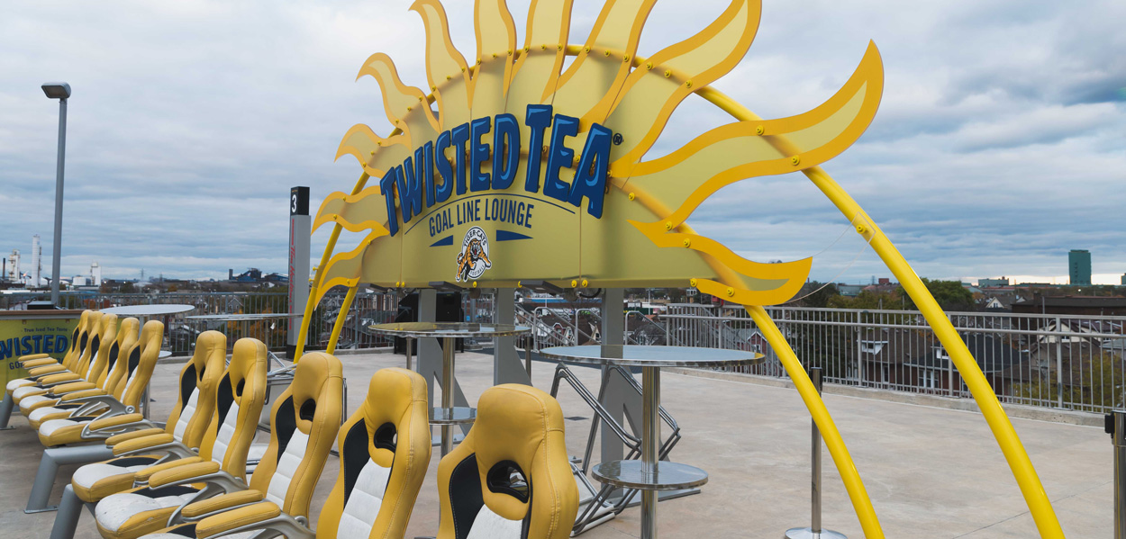 An outdoor Twisted Tea Goal Line Lounge features a large sun-shaped sign, a row of yellow and white stadium seats, and small tables, overlooking a city skyline under a cloudy sky.