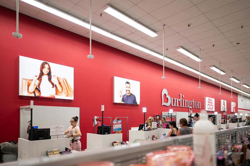 The checkout area of a Burlington store features a red wall with the logo, promotional images, and customers at registers.